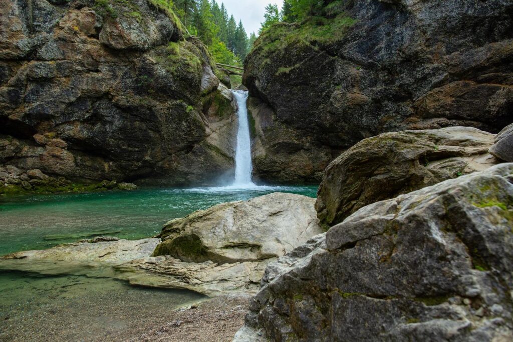 Klarer Wasserfall in einer Felsklamm mit türkisfarbenem Becken und großen Felsblöcken im Vordergrund.