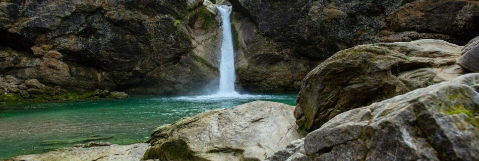 Klarer Wasserfall in einer Felsklamm mit türkisfarbenem Becken und großen Felsblöcken im Vordergrund.