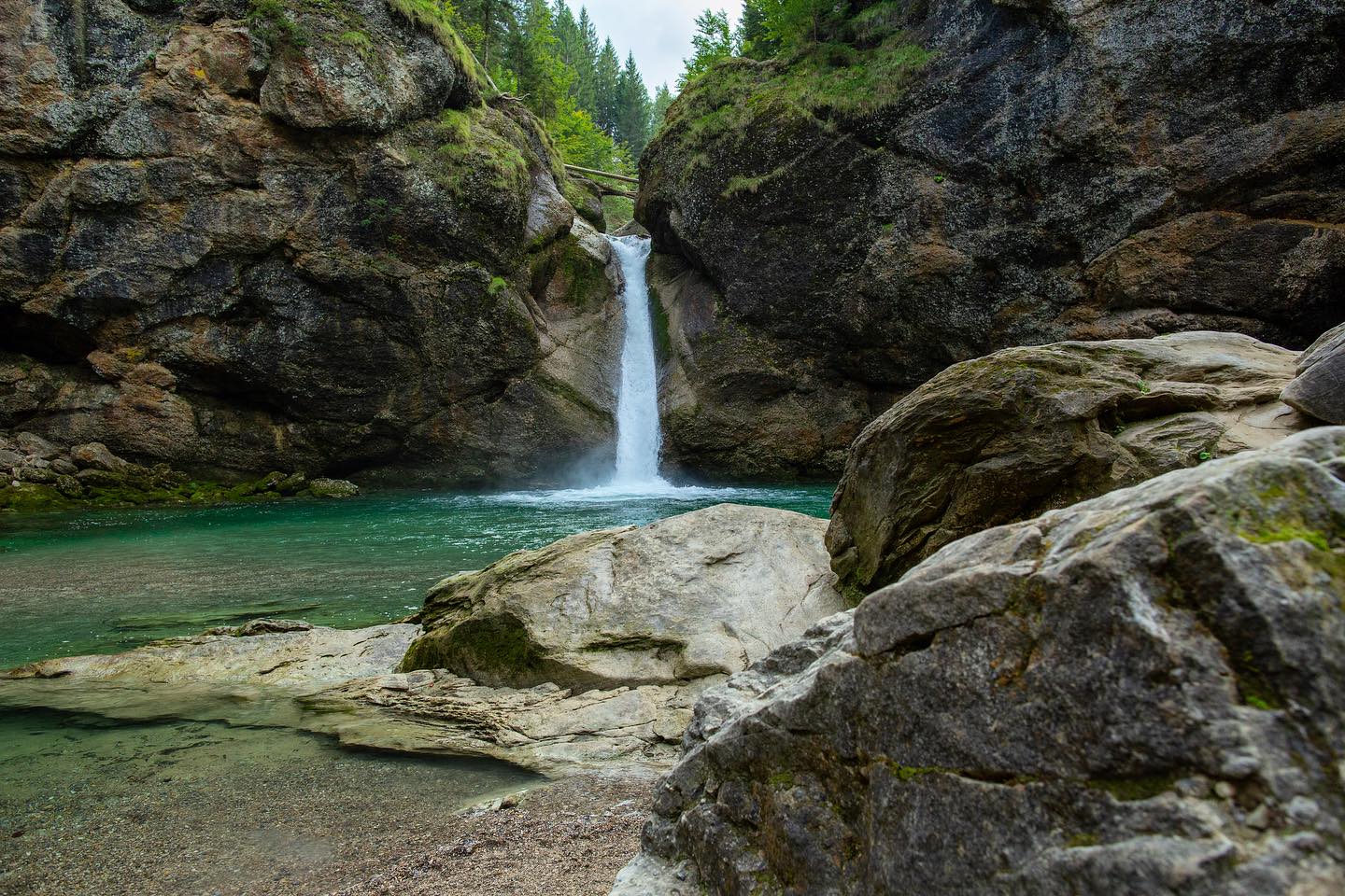 Klarer Wasserfall in einer Felsklamm mit türkisfarbenem Becken und großen Felsblöcken im Vordergrund.