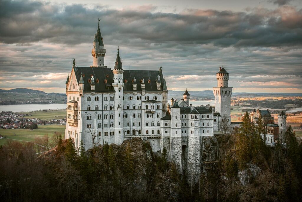 Schloss Neuschwanstein im warmen Abendlicht, umgeben von bewaldeten Hängen und weitem Alpenvorland unter dramatischer Wolkendecke.